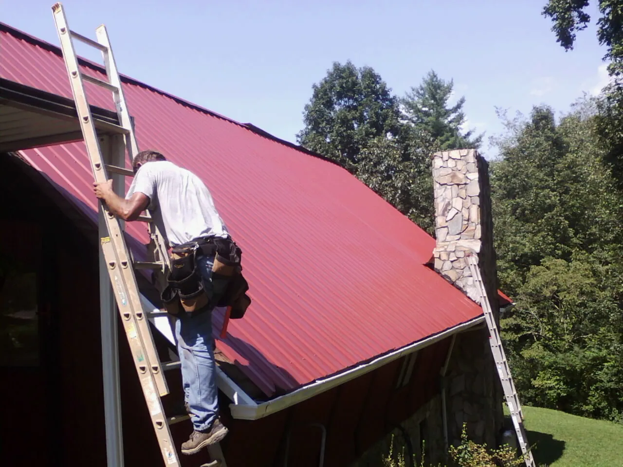Roofer on a ladder beside a red metal roof and stone chimney — Soffit & Fascia Repair by Roofing Brothers in Mcdonough
